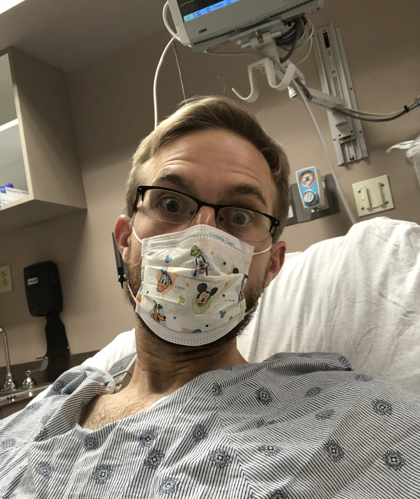 A person wearing glasses and a patterned medical mask is lying in a hospital bed, looking at the camera. They are dressed in a hospital gown. Medical equipment is visible in the background.