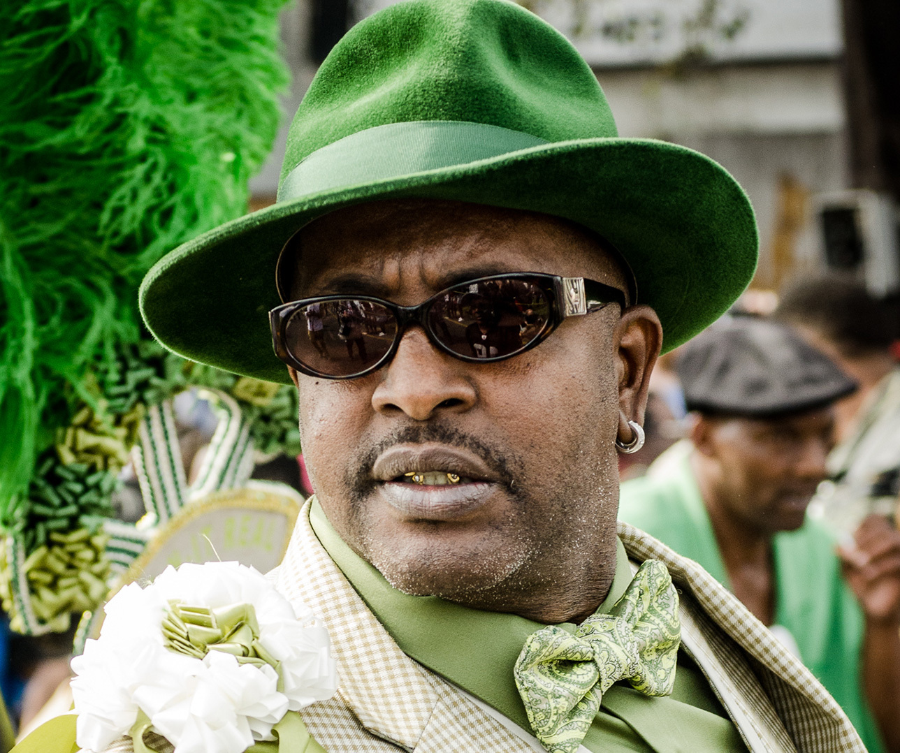 A man in a green suit, hat, and bow tie, wearing dark sunglasses, participates in a public event. He has a white flower on his lapel and is part of a festive, colorful crowd.