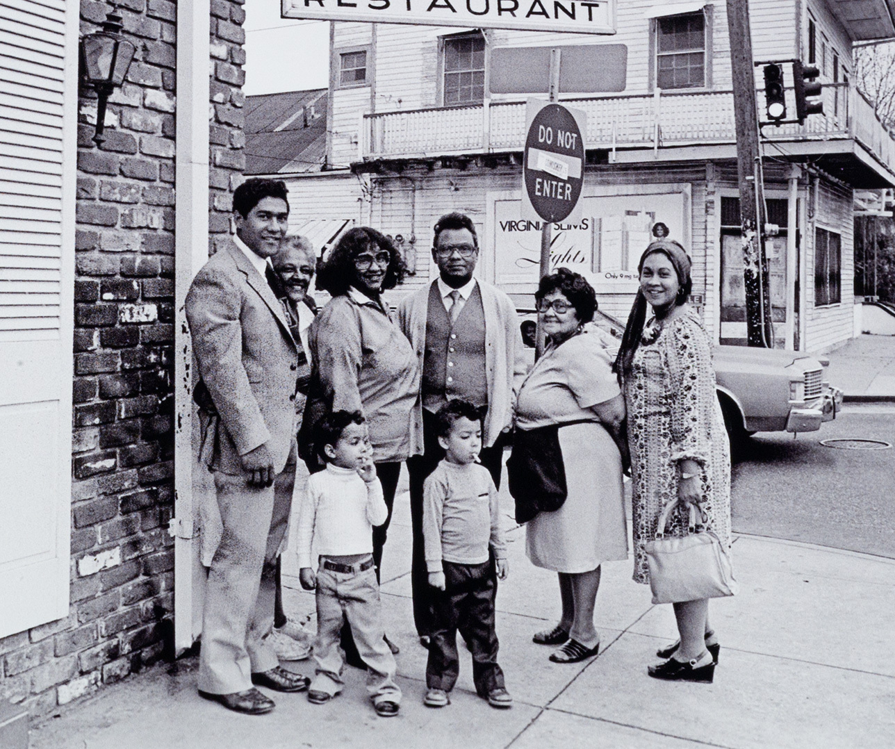 A group of eight people, including three children, pose on a street corner in front of a restaurant and a Do Not Enter sign. They are dressed in vintage attire, and a building is visible in the background.