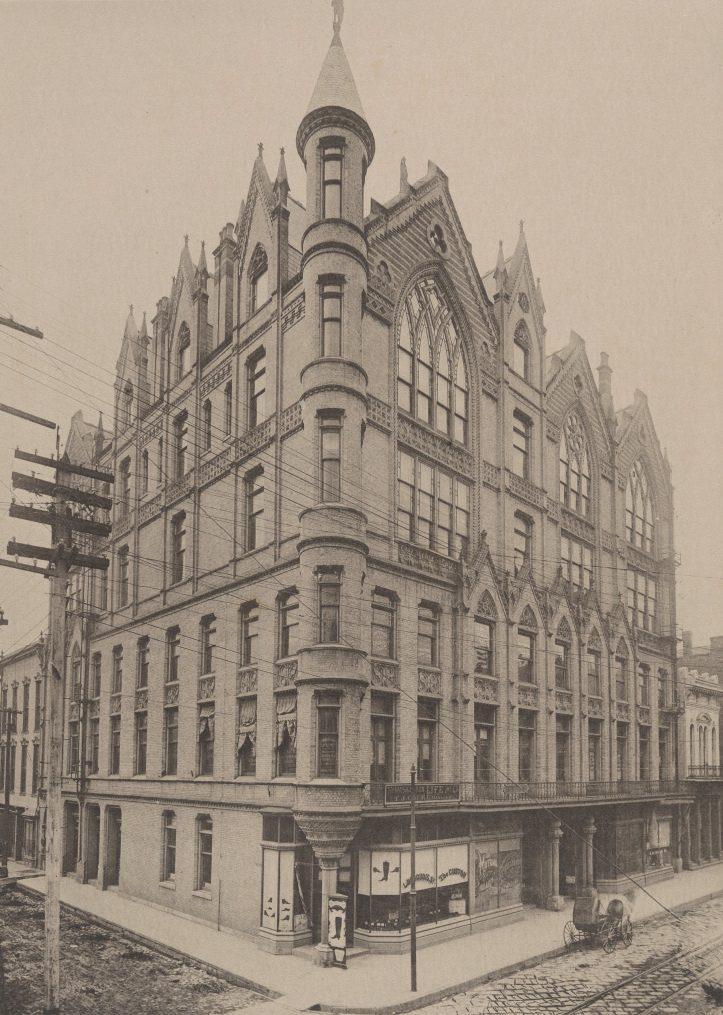 A historic, multi-story Gothic Revival building with pointed arches and ornate detailing. The corner structure features towers and large windows. A horse-drawn cart is parked on the cobblestone street nearby.