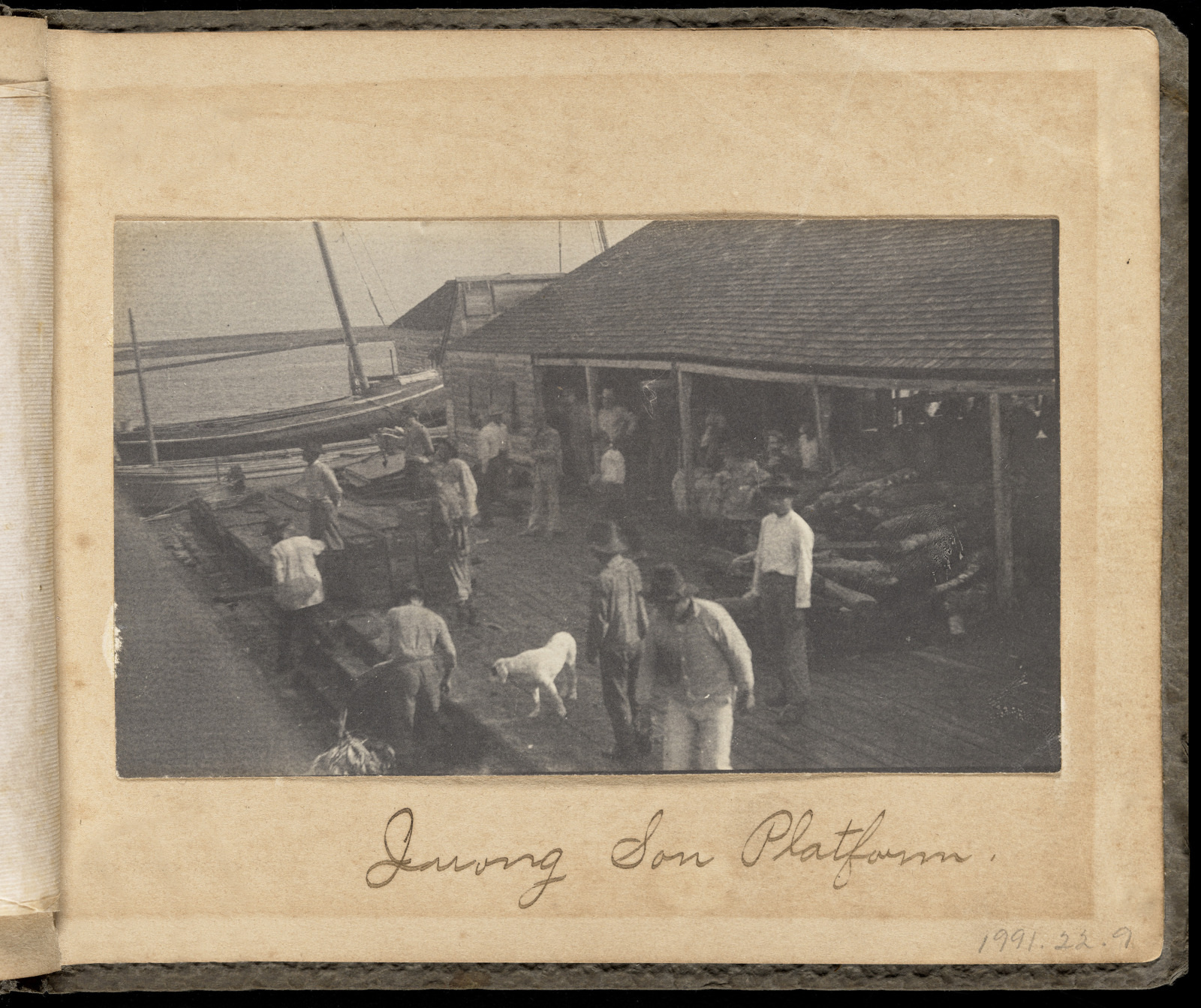 A vintage photo shows a group of people on a dock beside a building and a boat. Some individuals are carrying items, and a dog stands near them. The scene is labeled Drying Son Platform.