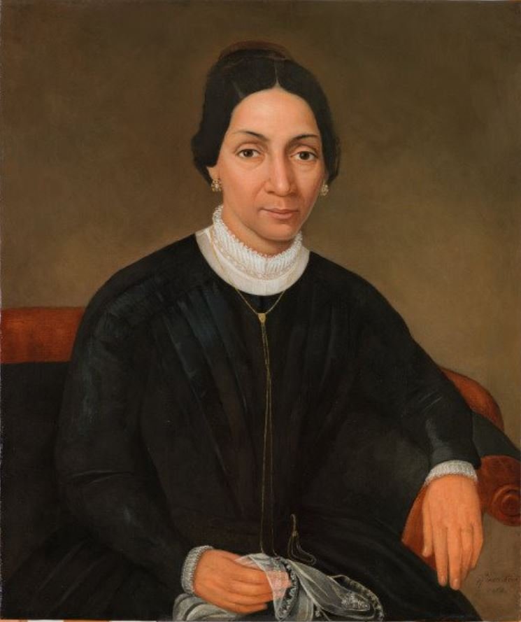 Portrait of a woman with dark hair, wearing a black dress with a white collar. She is seated, holding a small object in her left hand, against a plain background. Her expression is calm and composed.