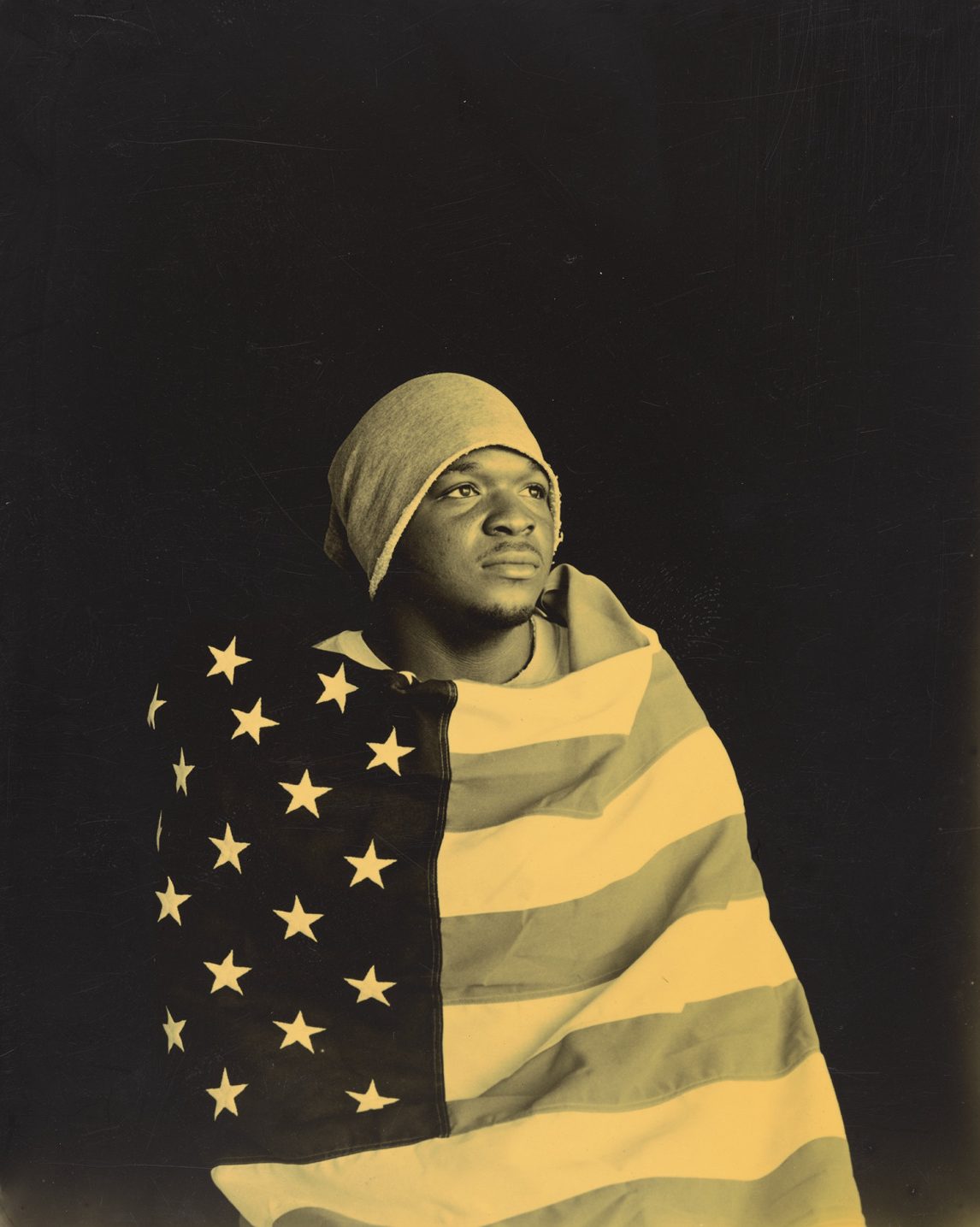 A sepia tone photograph shows a young incarcerated black man looking into the distance. He is wearing a beanie hat and is draped in an American flag.