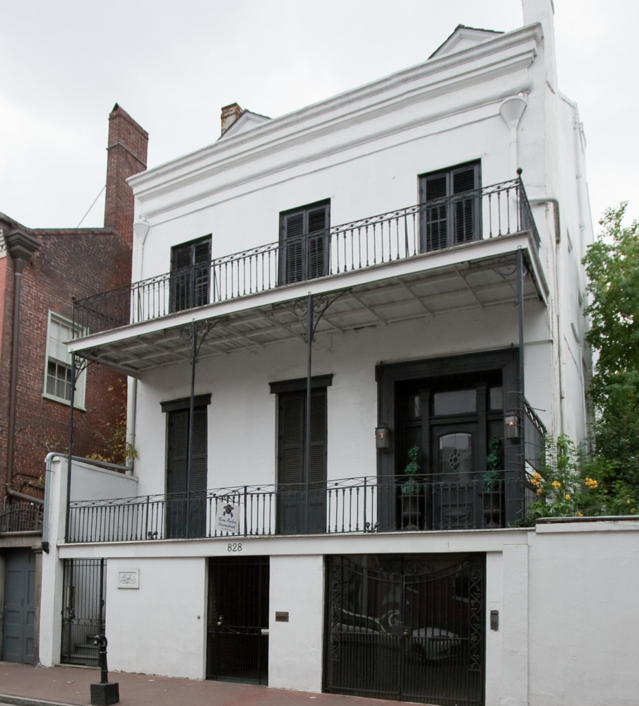 A three-story white building with black shutters and two balconies featuring wrought iron railings. A tree is visible on the right side, and the structure is nestled between brick buildings on a city street.
