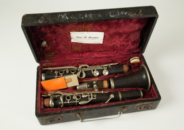 An open black case with red velvet lining containing a disassembled clarinet with silver keys. A cork grease container and an orange reed holder are also visible inside the case.