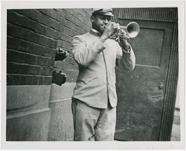 A man in a uniform plays a trumpet while standing next to a brick wall. He wears a cap and faces the right side of the image. Theres a door in the background. The photo is in black and white.