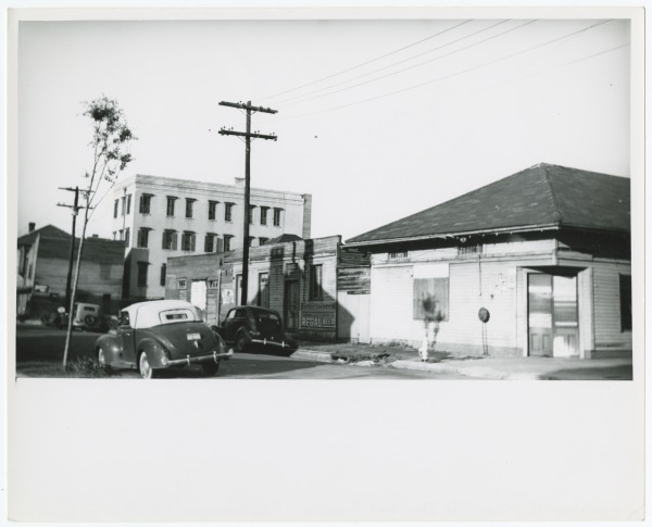 Black and white photo of a street scene with old-fashioned cars parked near wooden buildings. A multi-story brick building stands in the background. Power lines and a tree are visible, capturing a vintage urban atmosphere.
