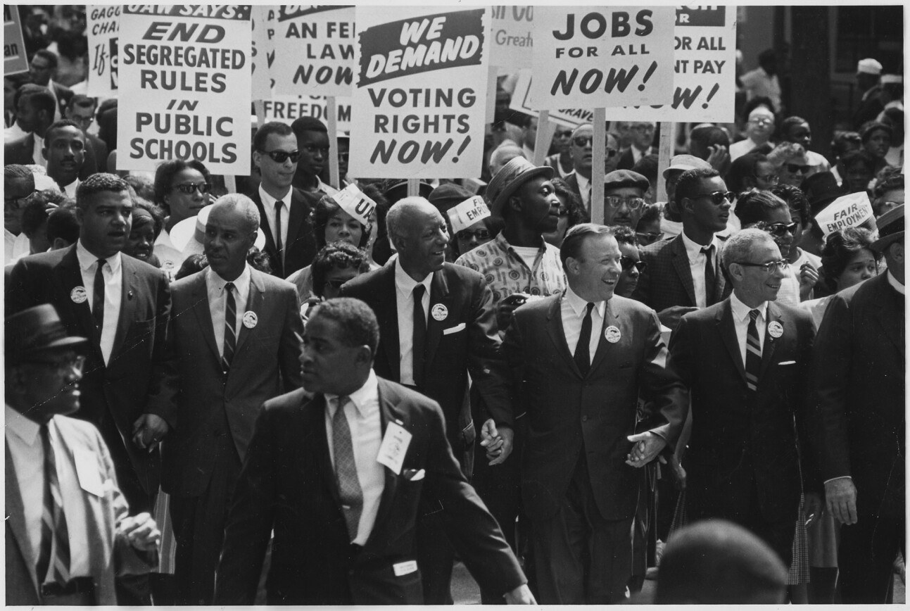 A black and white photo of a large group of people marching in a protest. They hold signs demanding voting rights, jobs, and an end to segregation. The expressions are determined as they walk together in solidarity.