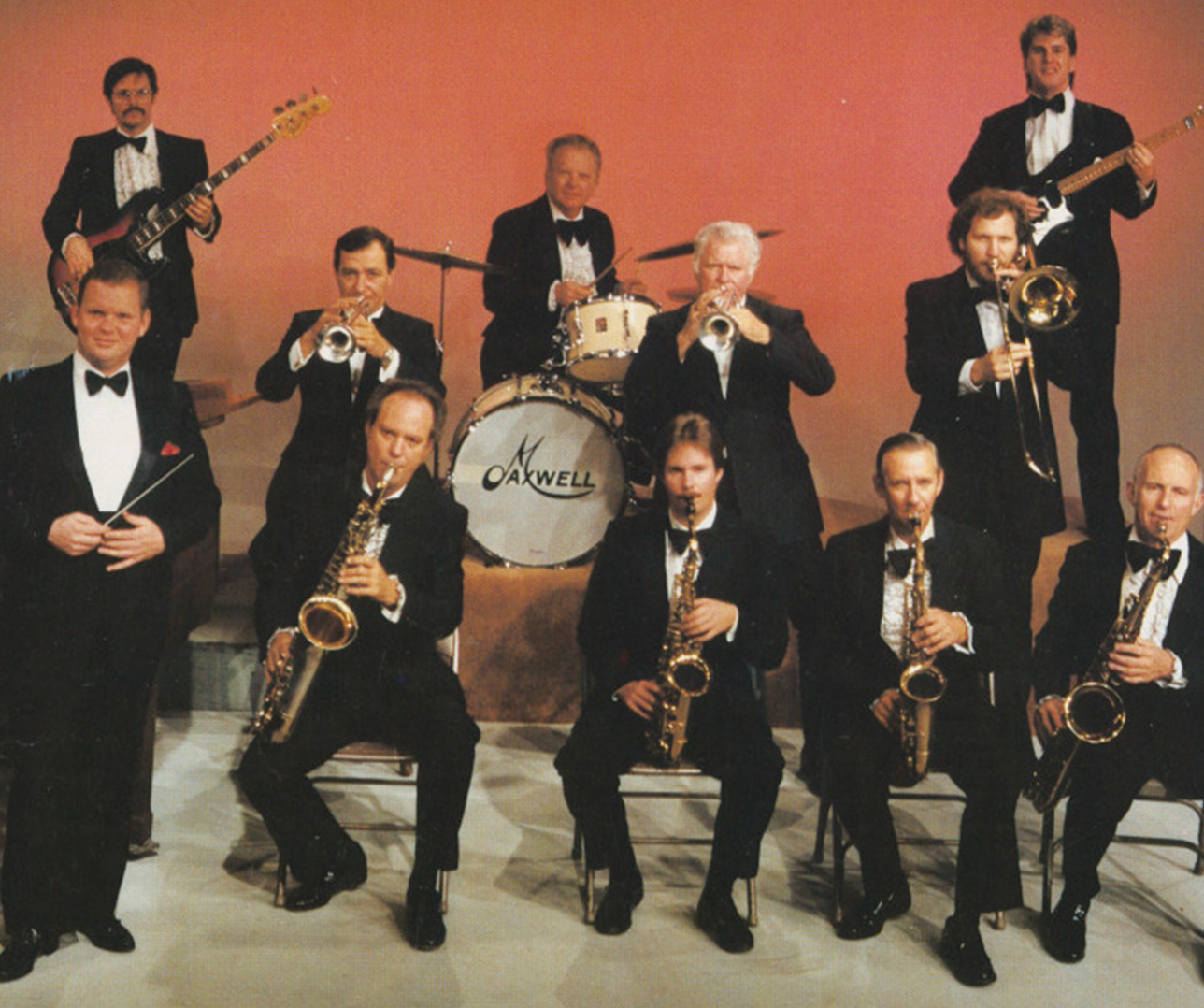 A big band ensemble in formal attire is performing on stage. Musicians are playing instruments like saxophones, trumpets, and a drum set. A conductor stands to the side. The backdrop is a warm, orange hue.