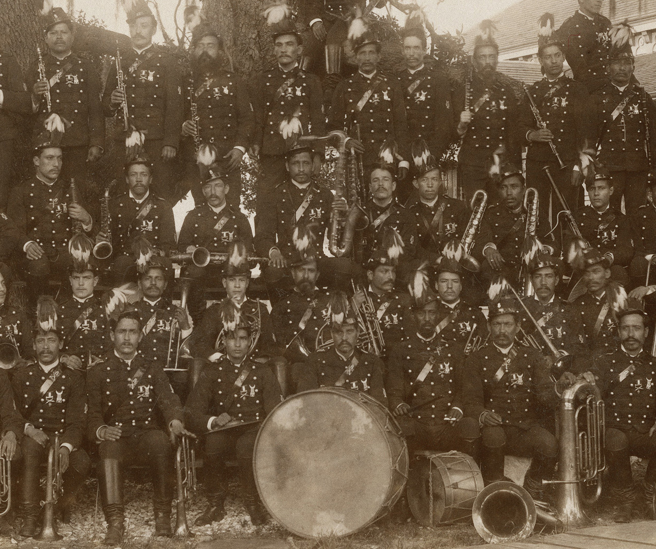 A sepia-toned vintage photograph of a large group of band members in uniform. They hold various musical instruments, including brass and percussion. The uniforms have decorative feathered helmets, and the group stands in rows outdoors.
