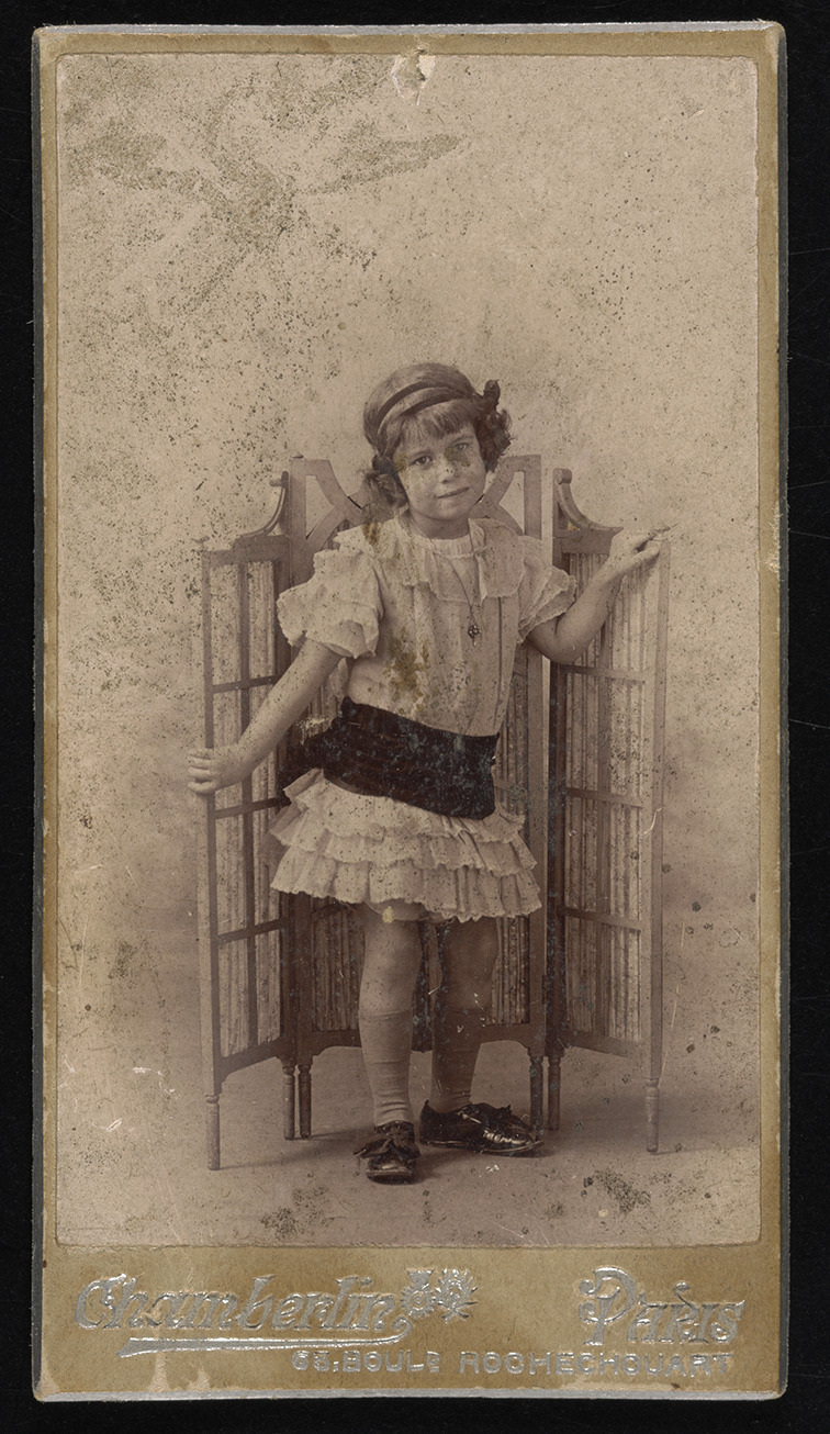 A vintage photograph of a young child wearing historical clothing, standing with one hand on a foldable wooden screen. The image has an old, faded appearance with visible wear and discoloration.