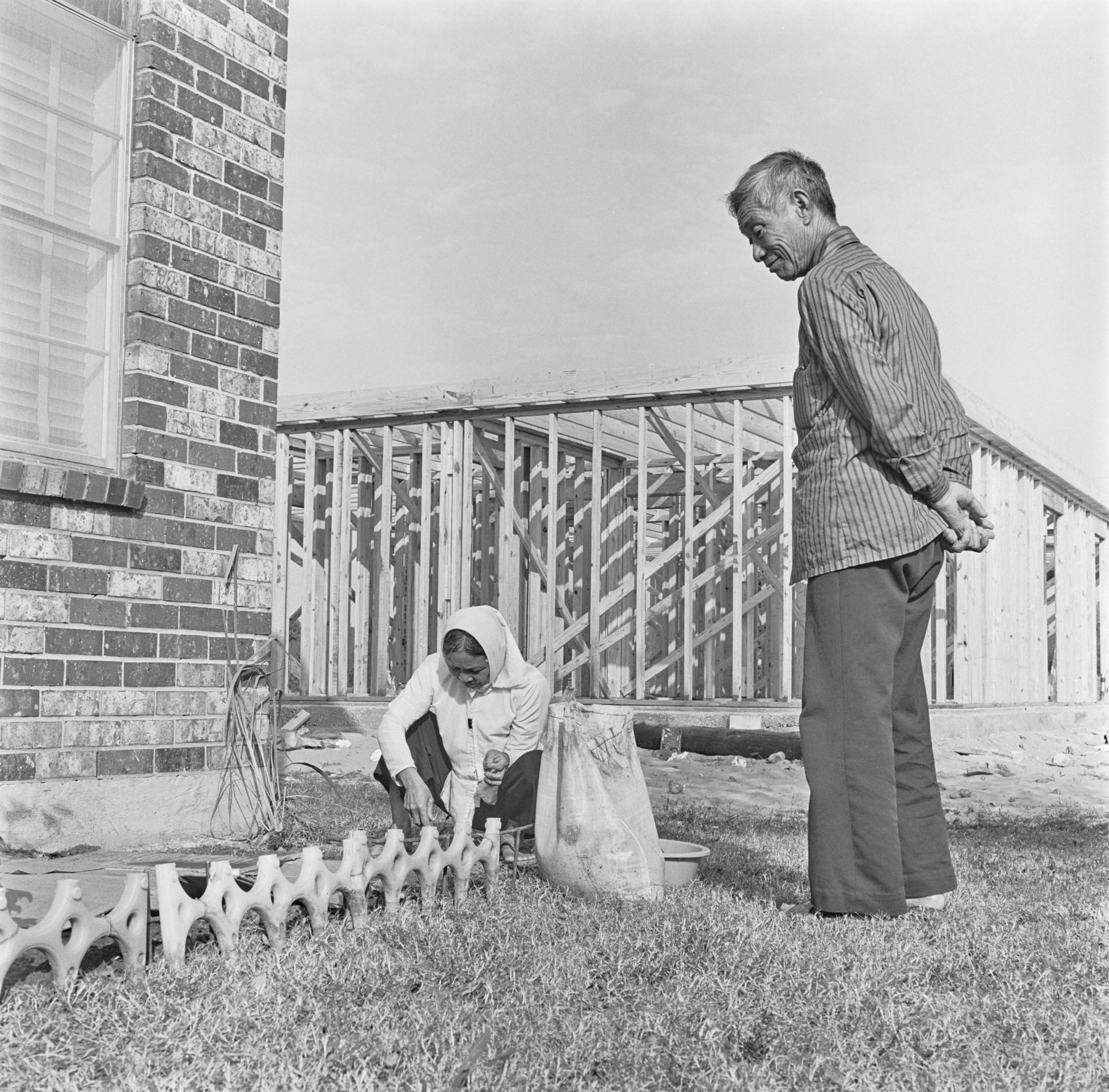 Two men in a yard with building materials. One man crouches, working near a brick wall, while the other stands nearby with his hands behind his back. A partially constructed house is in the background.