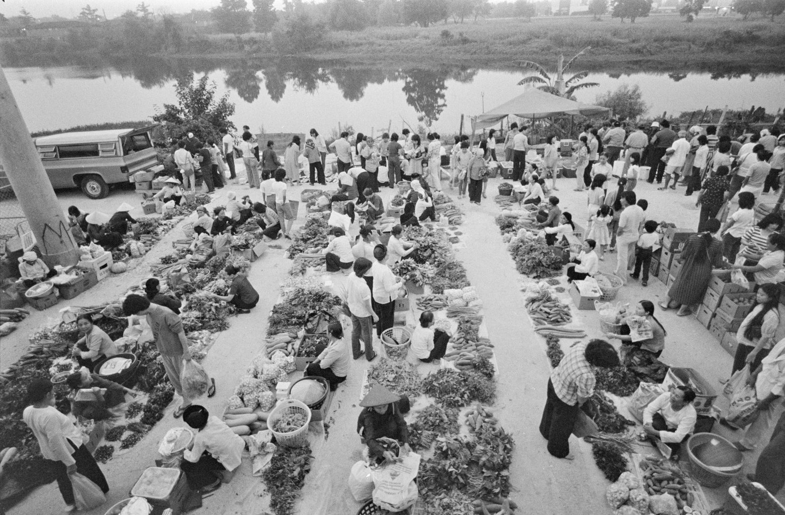 A vintage black and white photo shows a bustling market scene from above, with hundreds of people buying and selling goods. A river can be seen in the background.