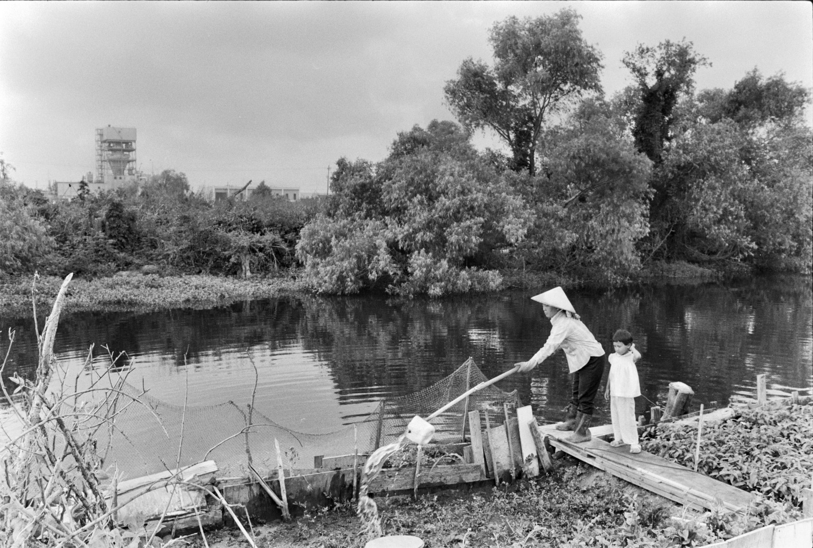 A small child looks on as a gardener uses a tao, or makeshift dipper, to water a garden plot in the Versailles community in 1983.