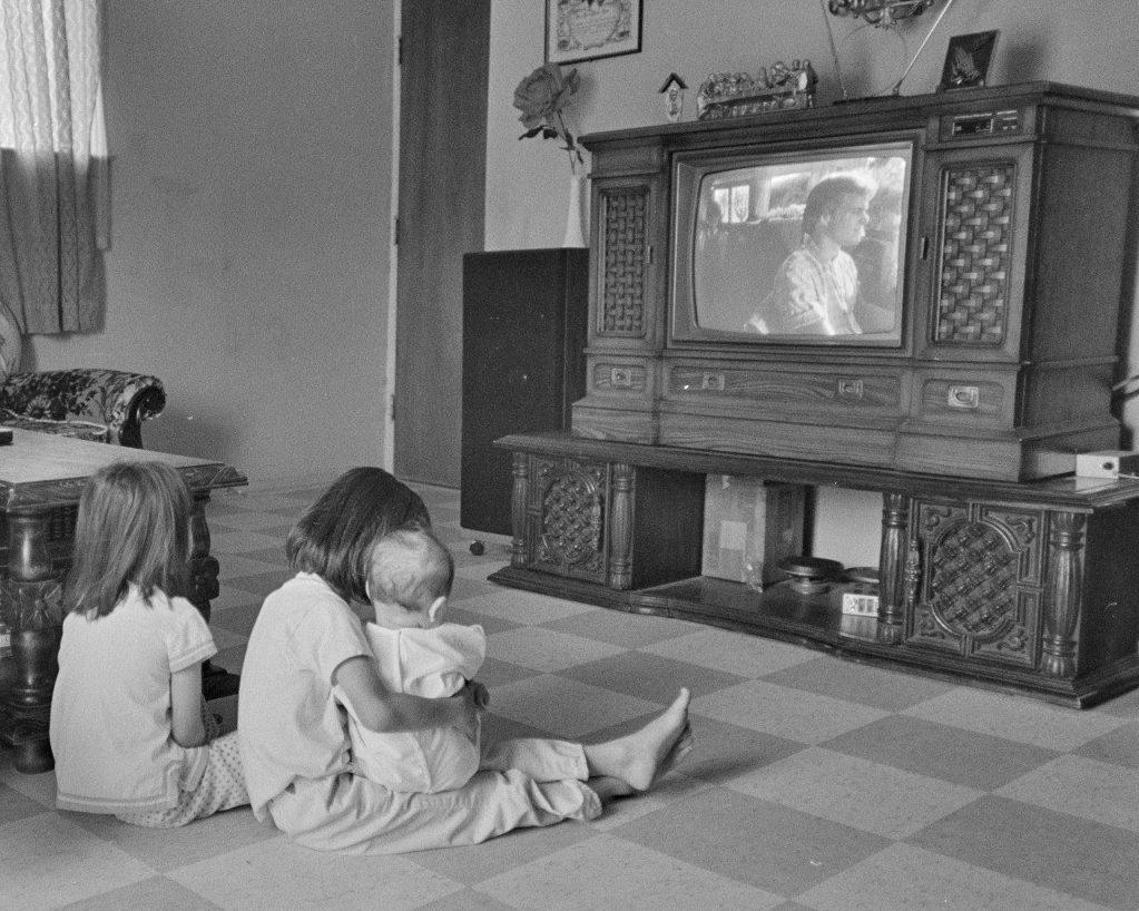 A black and white photo shows three children sitting on a tiled floor, watching a wooden console TV. One child holds a baby in their lap, while another sits nearby. The room is modestly furnished, with light coming through a window on the left.