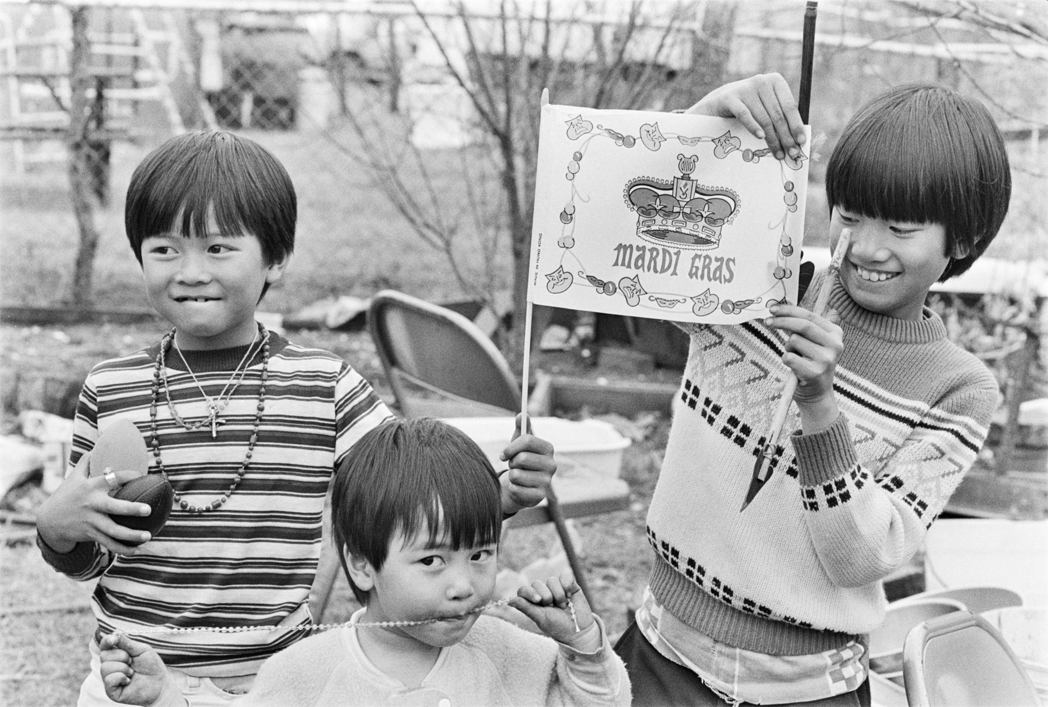A vintage black and white photo shows three Vietnamese children smiling to the camera while holding up Mardi Gras souvenirs including a flag.