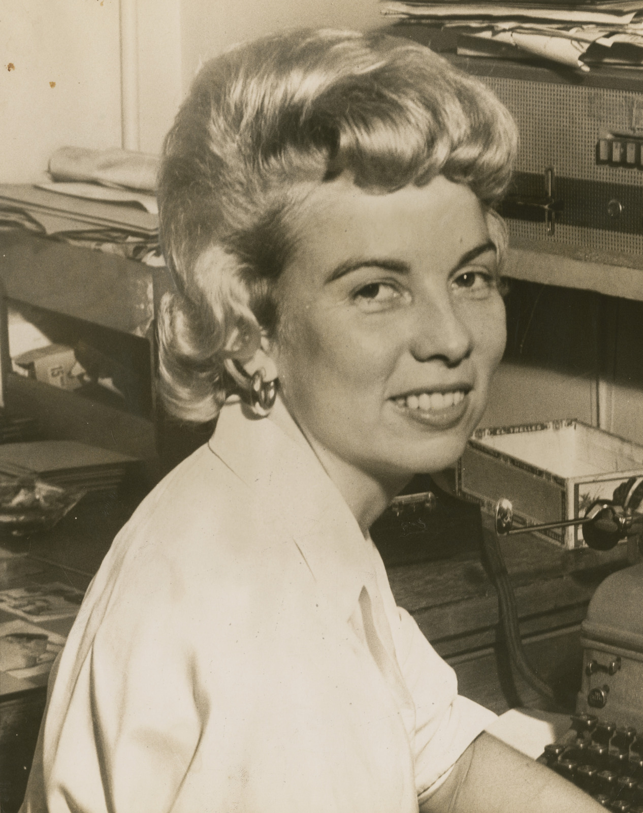 A woman with styled short hair and wearing a white blouse is sitting at a desk, smiling at the camera. The background shows papers, a typewriter, and an organized desk space.