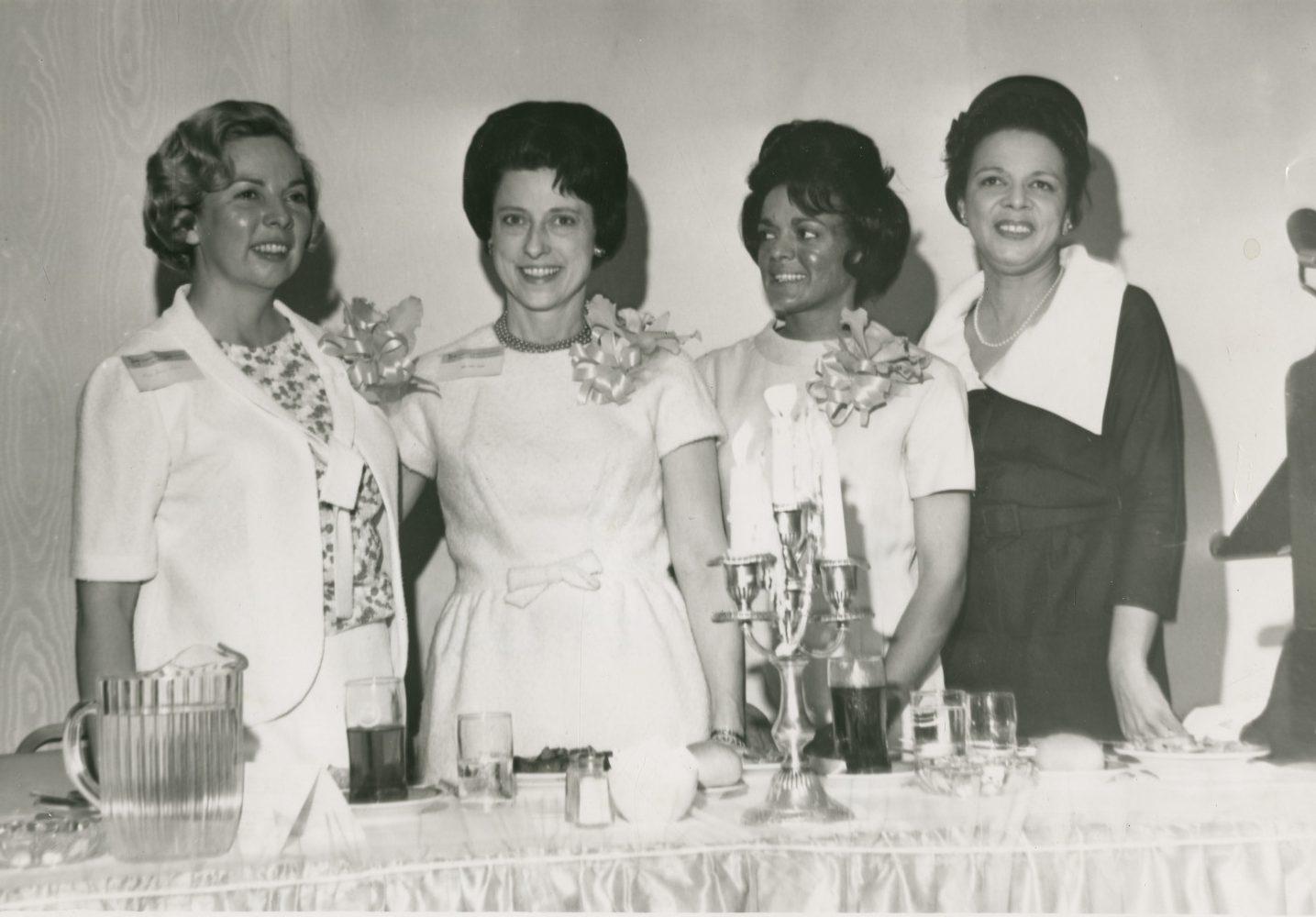 Four women stand behind a table set with a tablecloth, glassware, and a candle holder. Three of them wear light-colored dresses with floral corsages, and one wears a dark dress with a white collar. They are smiling and posed for a photograph.