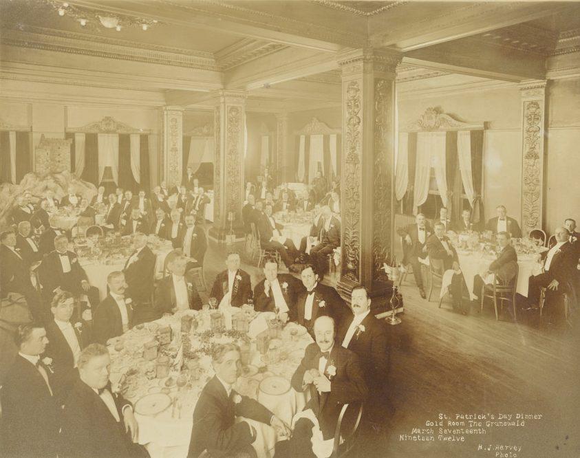 A sepia-toned photograph of a formal St. Patricks Day dinner. Numerous men in tuxedos sit at round tables in a grand hall with ornate columns and drapes. The scene conveys a sense of elegance and celebration.
