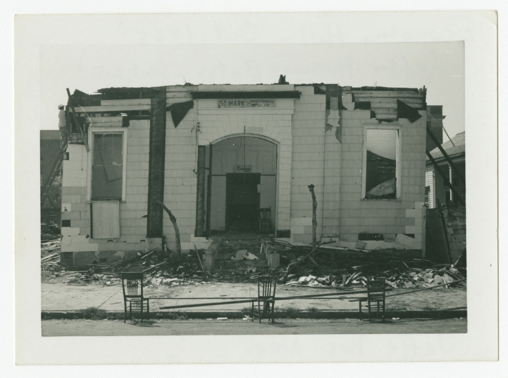 A black and white photo of a damaged building with missing roof and walls. Debris is scattered on the ground. The sign above the entrance reads BEER - SOFT DRINKS. Two empty chairs are on the pavement in front.
