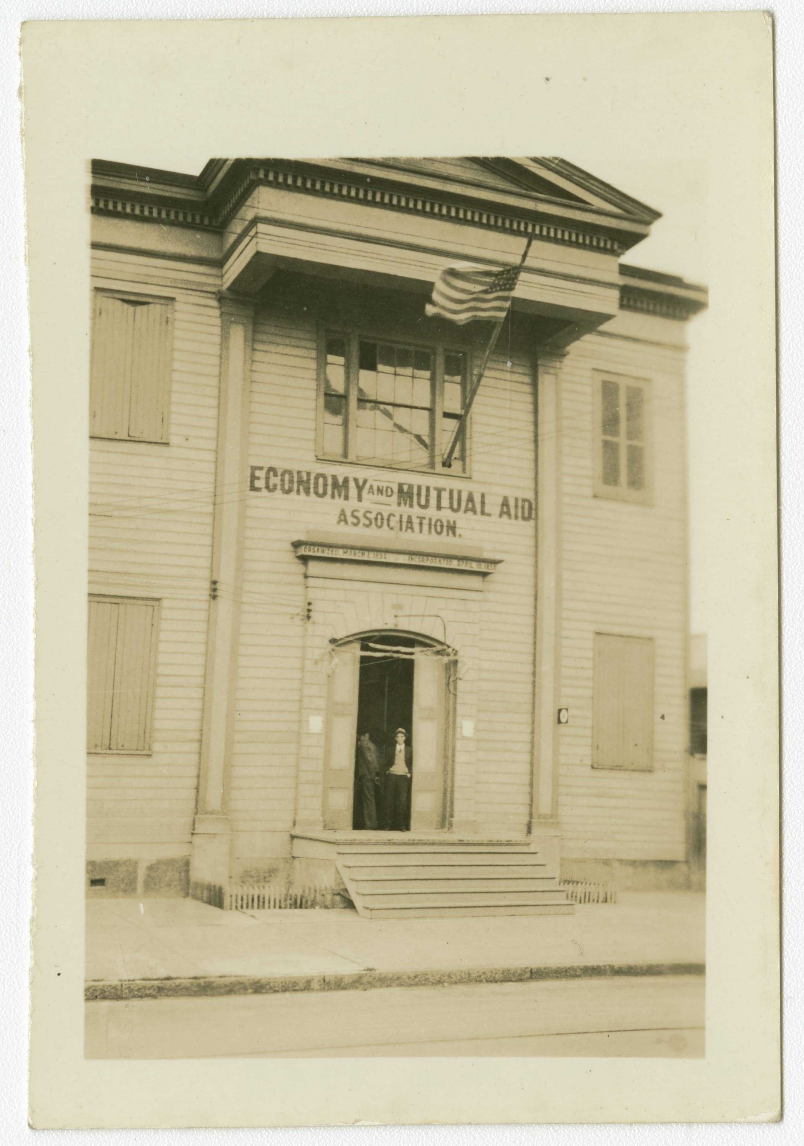 A black-and-white photo of a two-story wooden building with a sign reading Economy and Mutual Aid Association. The entrance has an open door, and two people are visible inside. An American flag hangs from the right side of the building.