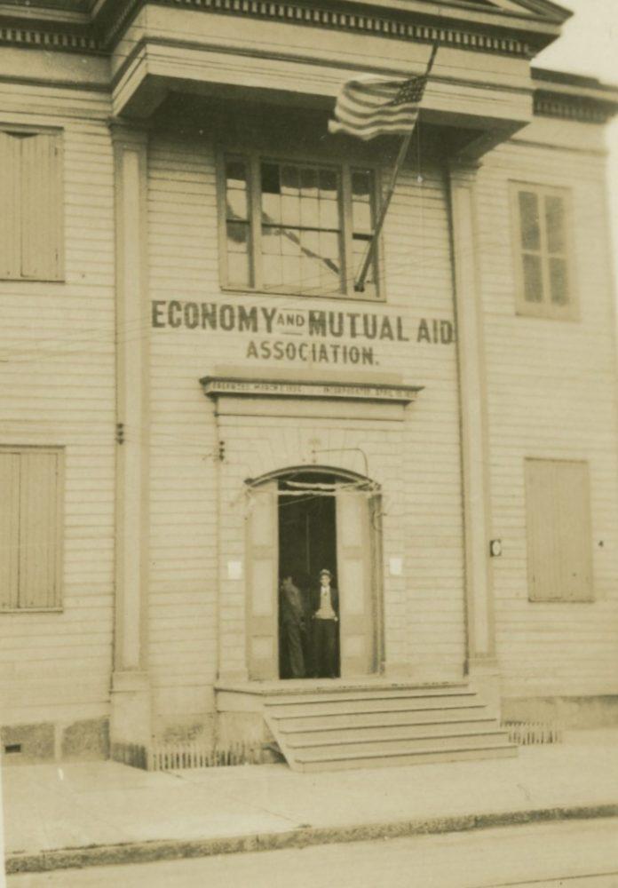 A vintage photograph of a wooden building with closed shutters. An American flag is hoisted above the entrance. A sign reads Economy and Mutual Aid Association. Two people stand in the doorway.