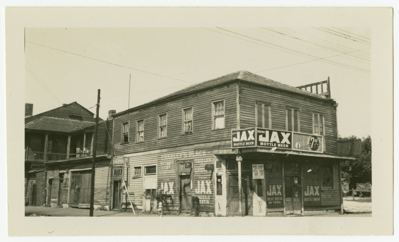 A vintage black-and-white photo of a two-story wooden building with signs advertising JAX Bottle Beer. The structure appears weathered, with a staircase on the right. Utility poles and wires are visible in the background.