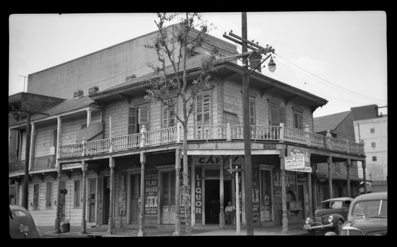 Black and white photo of a two-story corner building with a wraparound balcony and signs for a cafe, liquor store, and theater. Vintage cars are parked on the street, and a lamppost is visible. The architecture appears weathered and historic.