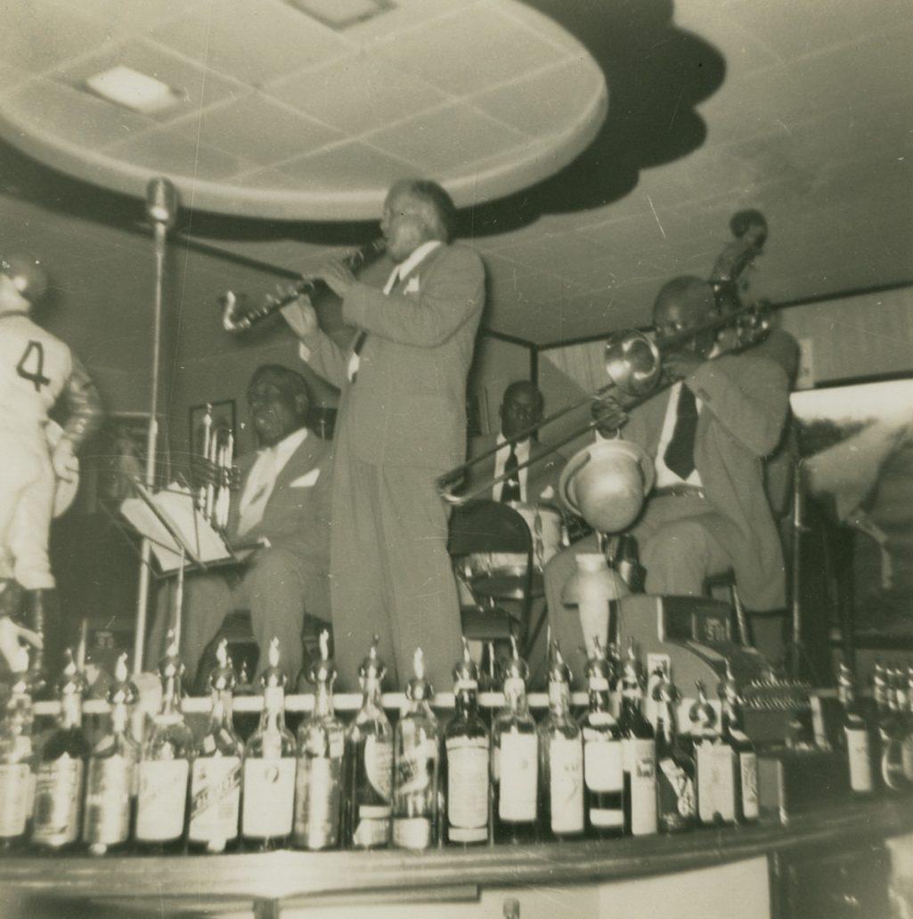 A black and white photo of a jazz band performing on stage. One musician is playing a clarinet, and another is on the trombone. Several bottles are lined up in the foreground, and a mannequin in a baseball uniform stands to the left.