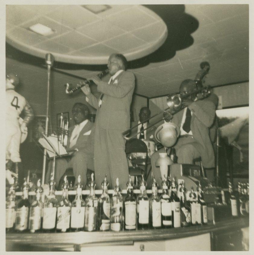 A vintage black and white photo of a jazz band performing on a stage. A saxophonist stands at the front while other musicians sit playing behind him. Numerous liquor bottles are lined up on the bar in the foreground.