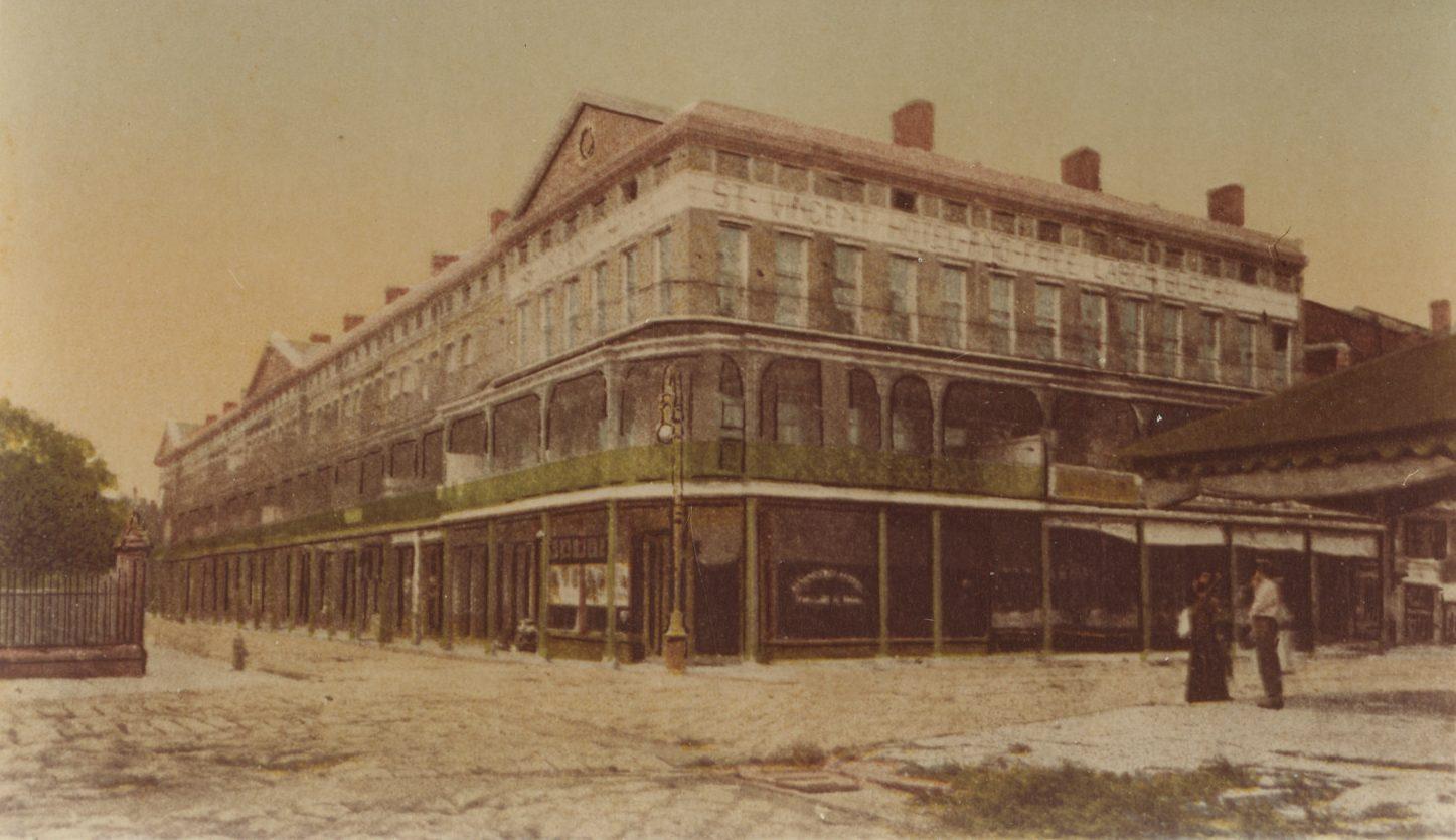 A vintage photograph of a large, multi-story building with long balconies and numerous windows, situated at a street corner. The street is cobblestoned, and two people stand talking nearby. The building has storefronts on the ground level.