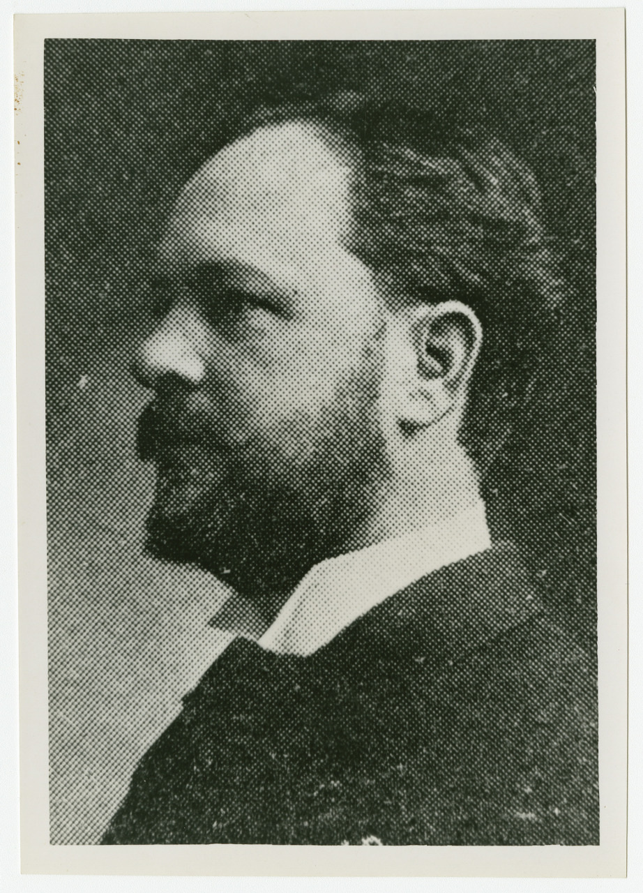 A black-and-white vintage photograph of a man with a beard and mustache, shown in profile with short hair, wearing a suit jacket and collared shirt. The image has a textured, dotted appearance typical of old prints.