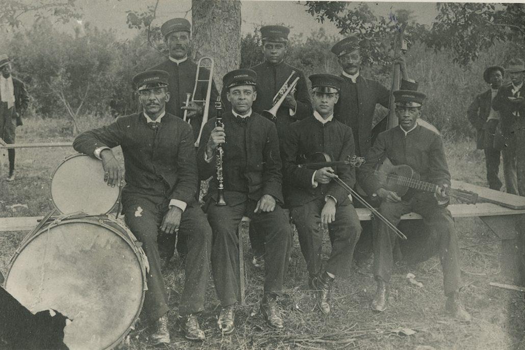 A vintage black and white photo of a band with seven men wearing uniforms and hats. They hold various musical instruments, including a trombone, clarinet, trumpet, violin, guitar, drum, and drumsticks, posed outdoors near trees.