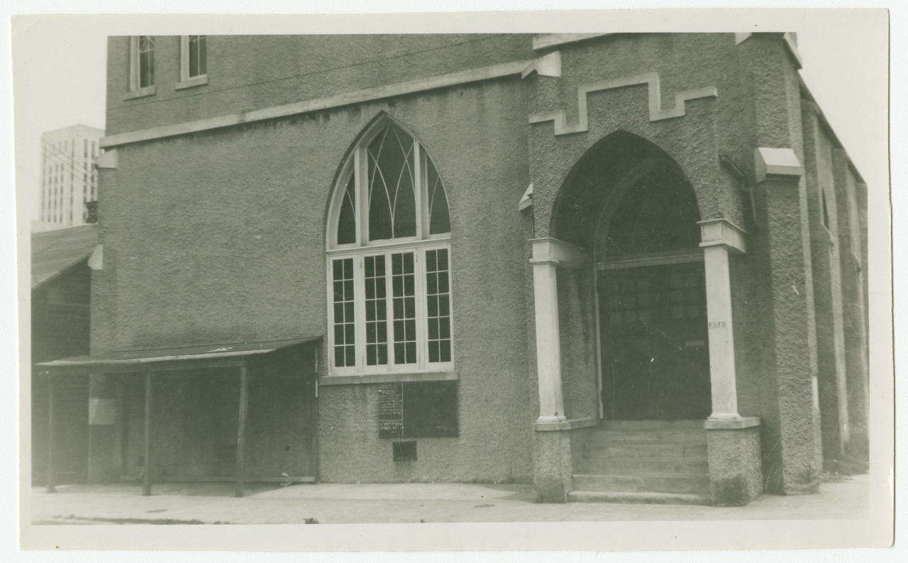 A vintage photograph of a church with a large arched entrance and a multi-paned arched window. The exterior is textured, and stairs lead up to the entrance. A small covered structure is attached to the side.
