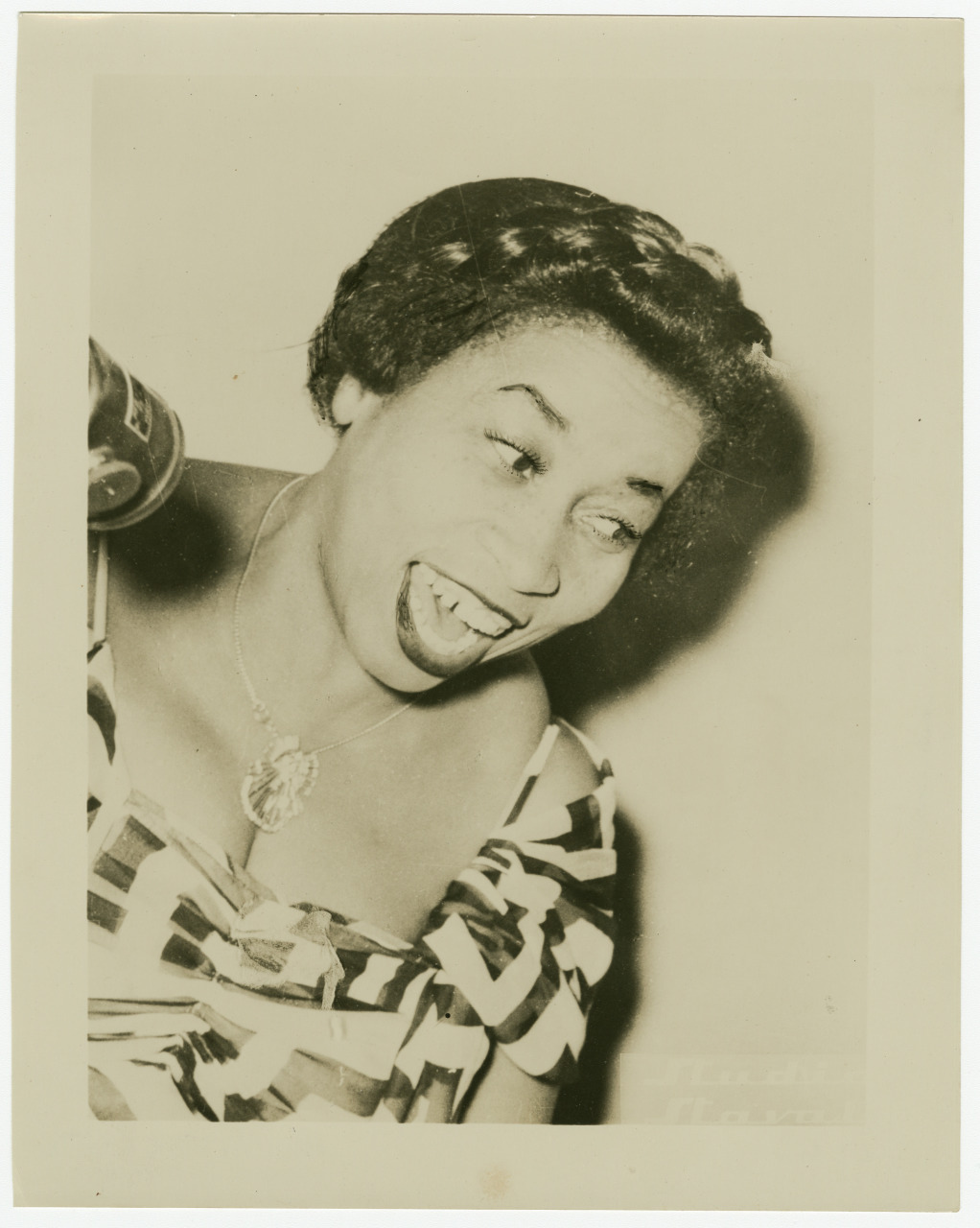A woman in a patterned dress sings into a vintage microphone. She has short curly hair and is wearing a necklace with a pendant. Her expression is animated and joyful. The image is in black and white.