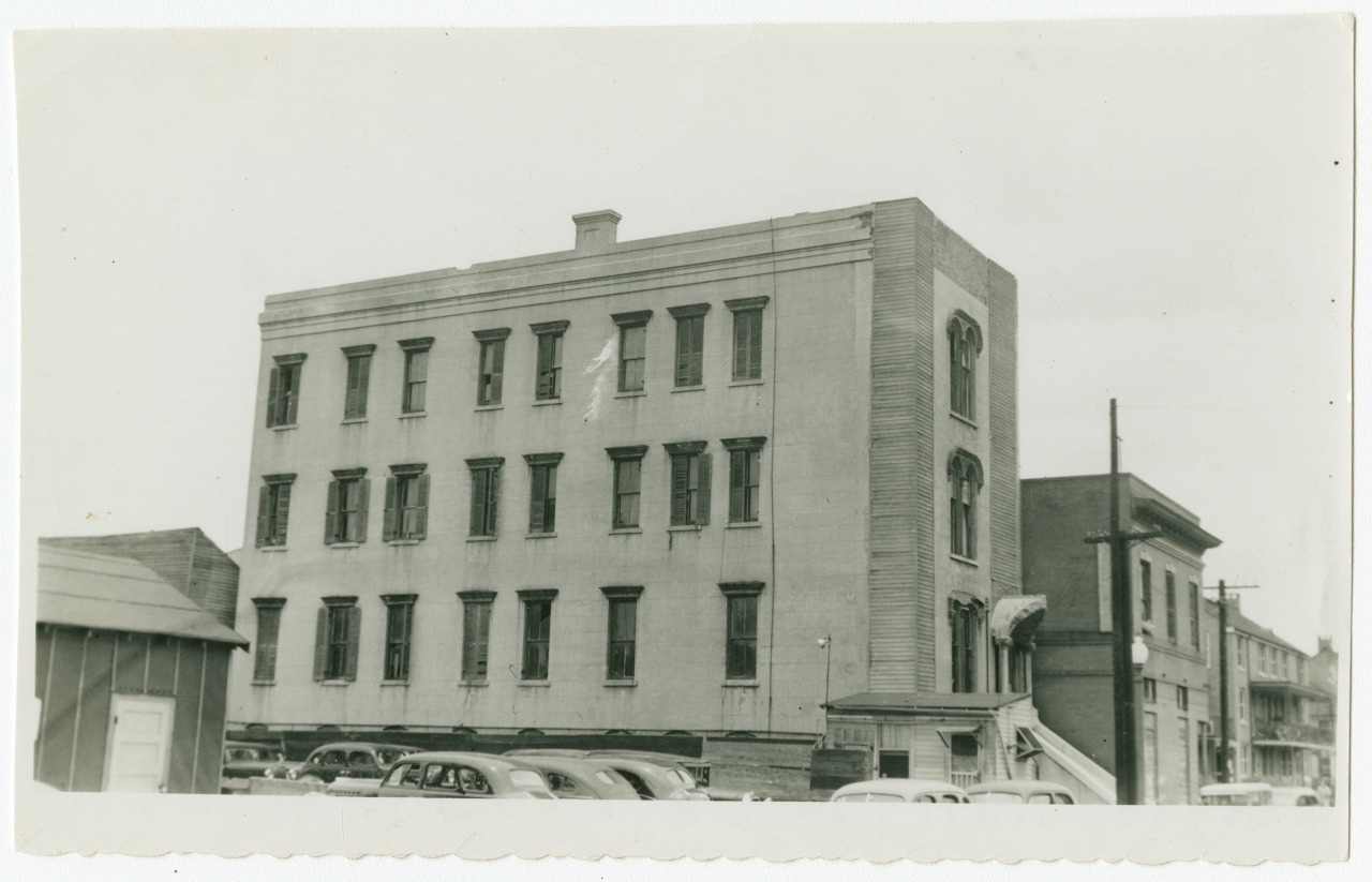 A vintage black and white photo of a three-story brick building with multiple windows. Several classic cars are parked in front, and adjacent structures are visible on a cloudy day.