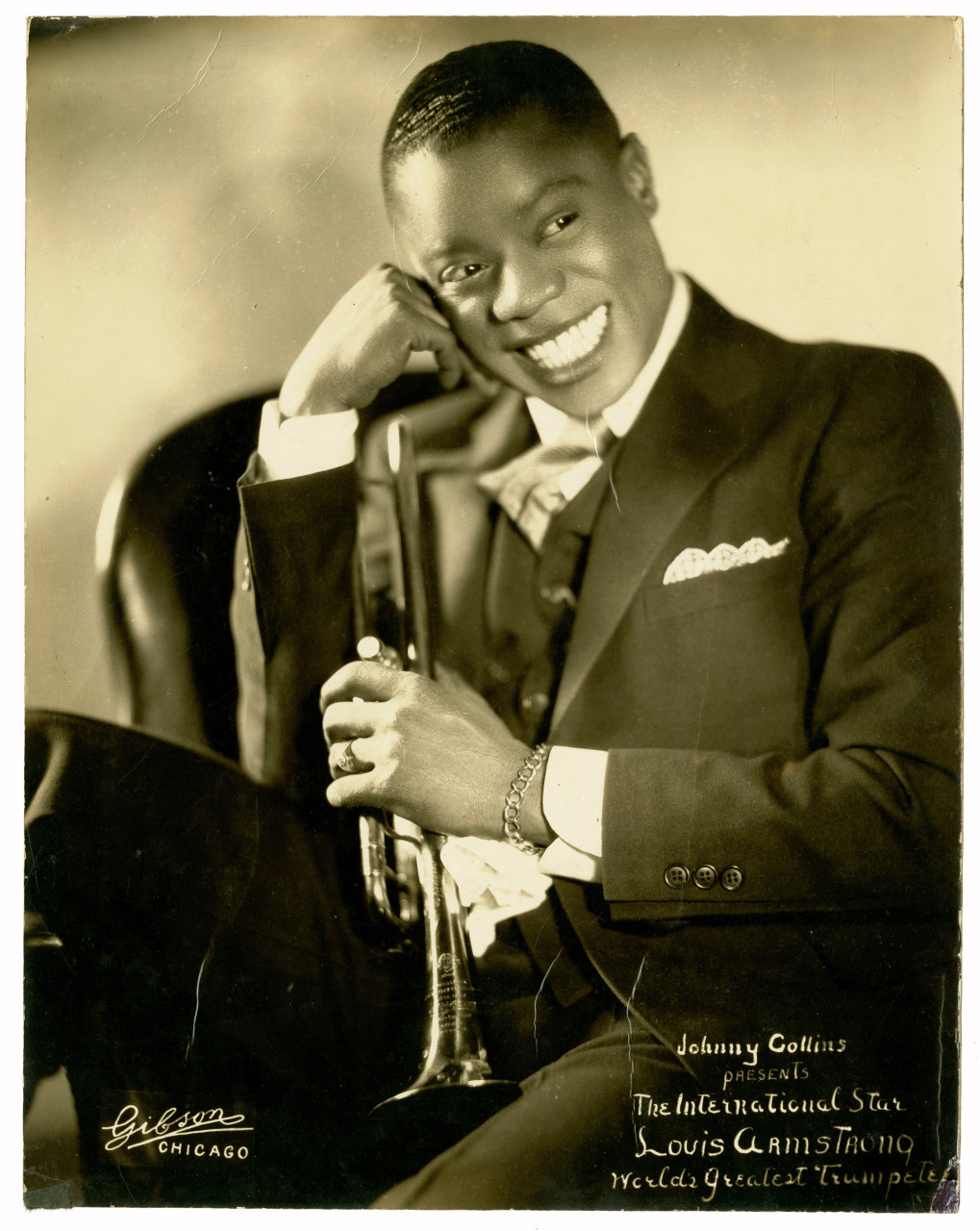 Vintage portrait of a smiling man in a suit, holding a trumpet. The text reads Johnny Collins presents the international star Louis Armstrong Worlds Greatest Trumpeter.