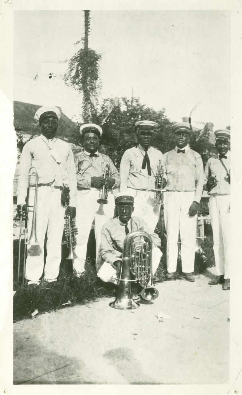 A vintage photo of six band members in uniforms and hats, holding brass instruments, including a trombone, trumpet, and tuba. One person is seated on the ground. They are outdoors with trees and buildings in the background.