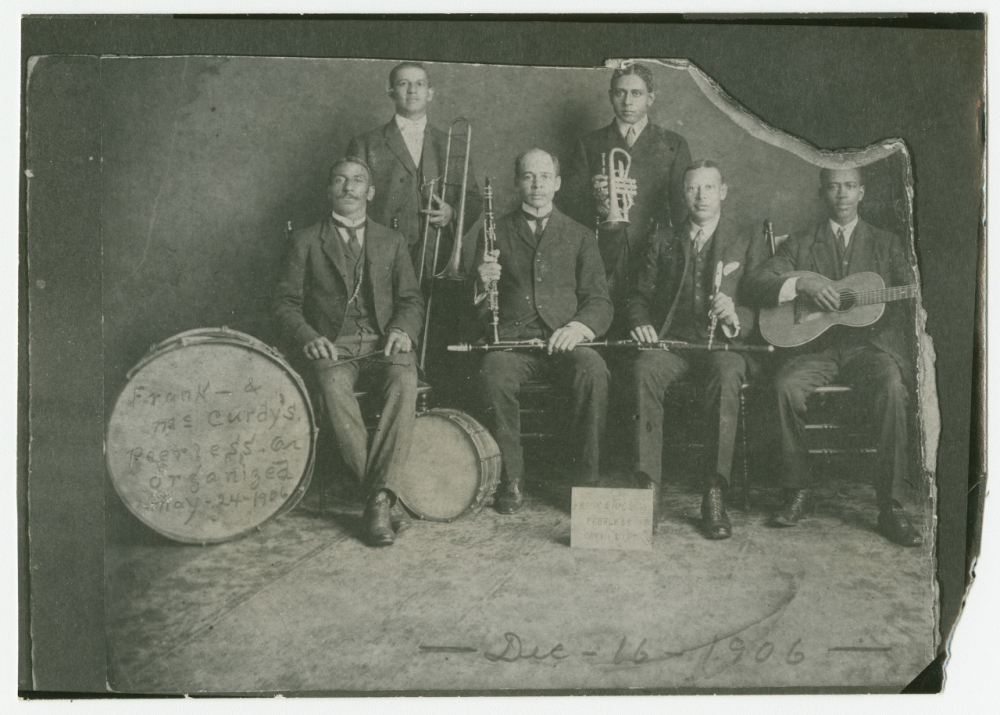 A vintage black and white photo from 1906 showing six men in suits holding musical instruments, including a trombone, cornet, clarinet, and guitar. A large bass drum is visible with handwriting. The photo has a worn and torn appearance.