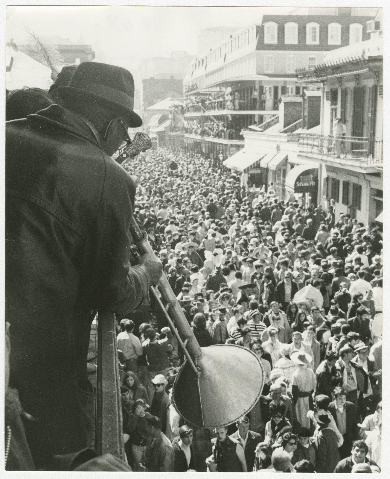A black-and-white photo of a crowded street scene during Mardi Gras in New Orleans. A musician plays a trombone on a balcony above, overlooking a dense crowd of people in the street below. Buildings with balconies line the street.