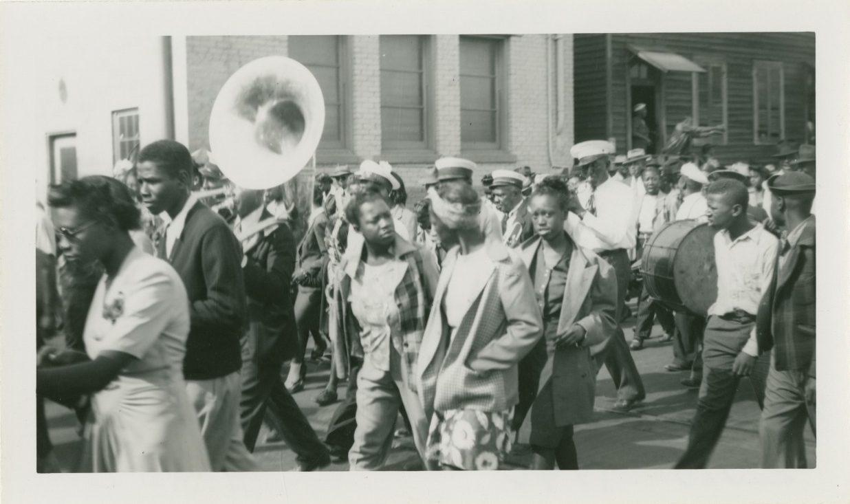 A black and white photo showing a street parade with musicians playing brass and percussion instruments. People walk alongside, wearing mid-20th century clothing. Buildings are visible in the background.