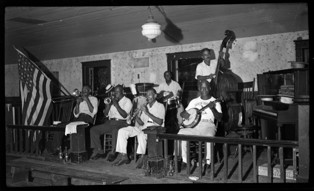 A black and white photo of a jazz band performing on stage. The musicians are playing various instruments including a trumpet, clarinet, trombone, banjo, and double bass. An American flag hangs on the side, and theres a vintage light fixture above.