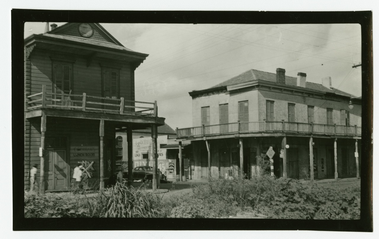 A black and white photograph of two old, wooden buildings on a street corner. One building has a balcony with a wooden railing, while the other features a wraparound porch. A vintage truck is parked nearby, and clouds fill the sky.