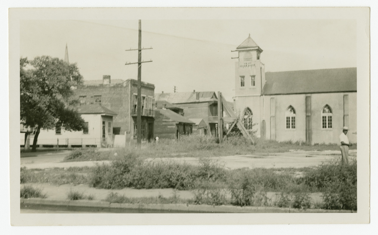 A vintage black and white photo of a vacant lot overgrown with weeds. In the background, there is a church with a bell tower and other surrounding buildings. A man in a hat stands on the right side, facing the scene.