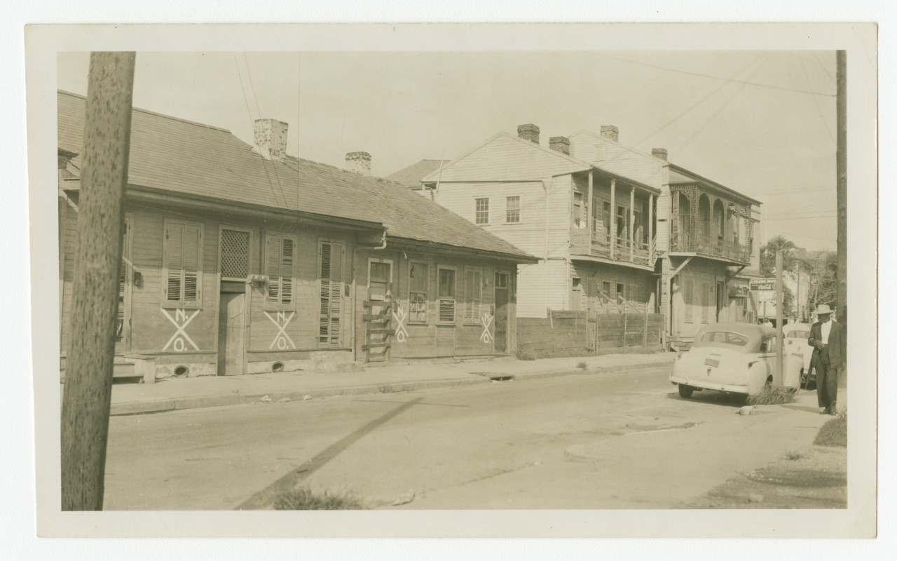 A black-and-white photo of a street with old wooden buildings on one side. Two-story structures feature balconies and shutters. A vintage car is parked on the road, and a few people stand nearby. Power lines crisscross above.