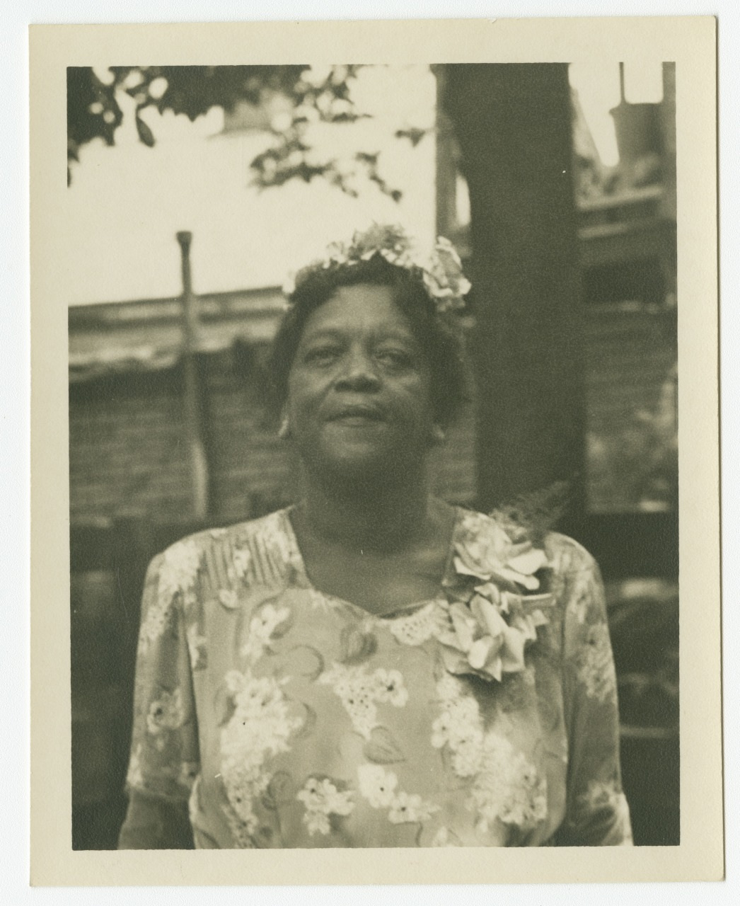 Black and white photo of a woman standing outdoors, wearing a floral dress and floral headpiece. Trees and a building are in the background.