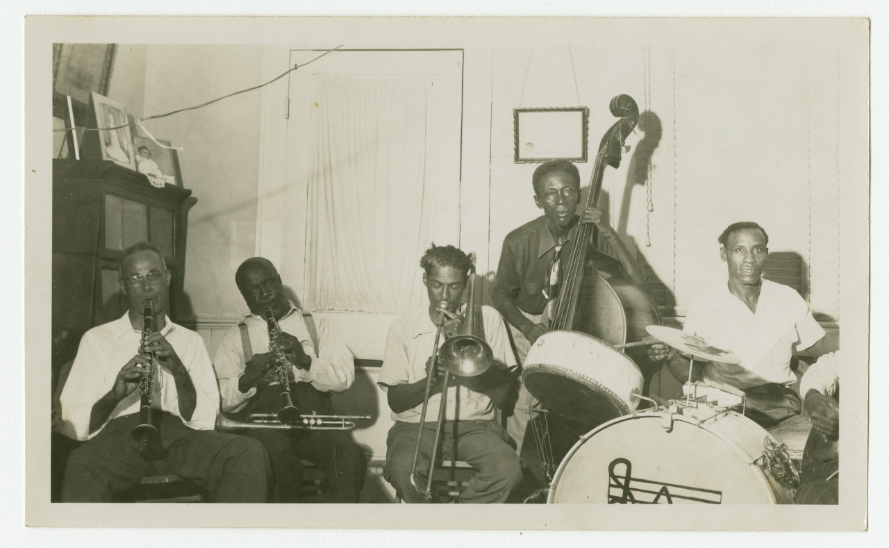 A vintage black and white photo of a jazz band with five musicians. They are playing various instruments, including clarinet, trumpet, trombone, double bass, and drums, in a room with a window and framed picture on the wall.