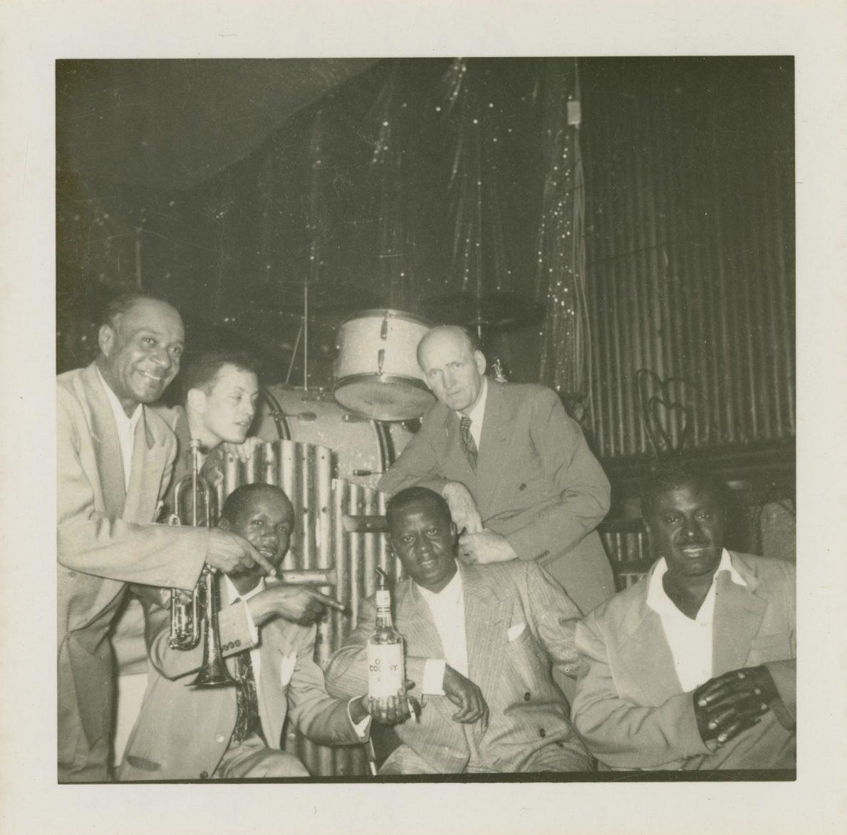 A group of six men pose together in a dimly lit room. Two men hold musical instruments, including a trumpet. One man holds a bottle. They are dressed in suits and appear to be enjoying themselves in a casual setting.