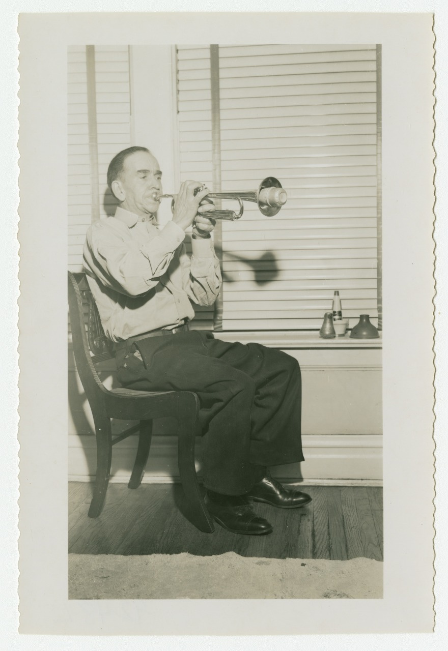 A man sitting on a chair, playing a trumpet. He faces a set of closed shutters, with two trumpet mutes resting on the windowsill beside him. The image is black and white and has a vintage appearance.