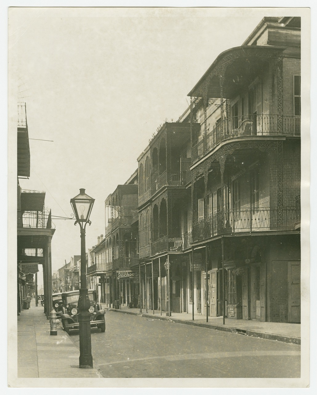 Black and white photo of a vintage street scene, featuring historic buildings with wrought iron balconies. A vintage car is parked along the sidewalk. Street lamps line the cobblestone street, creating an early 20th-century atmosphere.